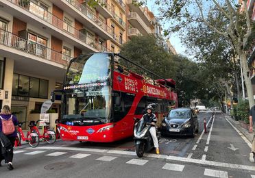 A bright red open-top hop-on hop-off bus in Barcelona, filled with tourists, with landmarks like Sagrada Família and Park Güell in the background under a sunny Mediterranean sky