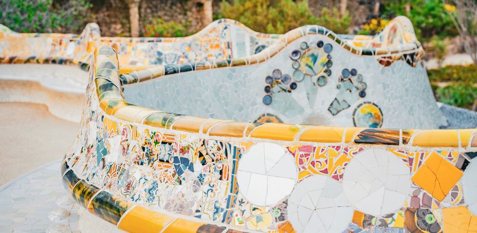 Close-up view of a colorful mosaic-covered bench in Park Güell, Barcelona, showcasing intricate tile patterns in yellow, orange, and white tones, with a blurred natural background featuring arches and greenery.